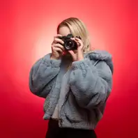 Eine junge Frau posiert stehend vor einer roten Fotostudiowand. Sie halt eine Fotokamera vor ihrem Gesicht